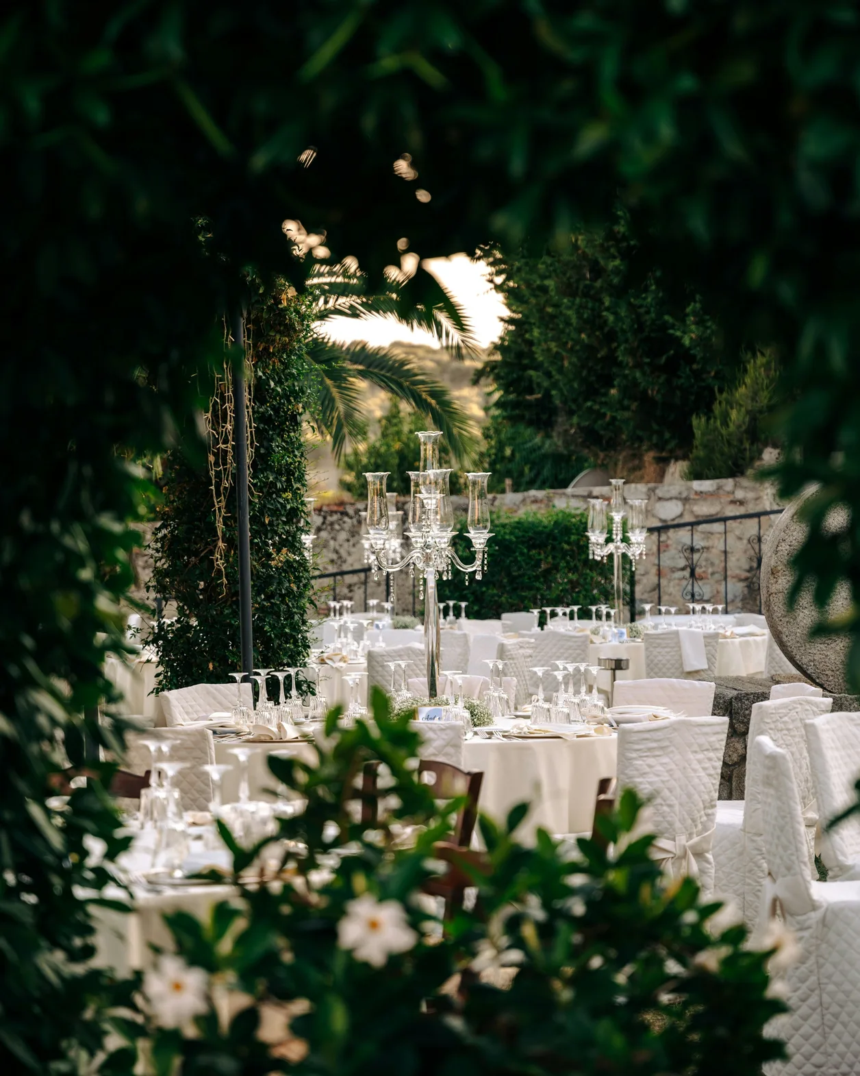 Garden dinner glimpsed through foliage, with tall candelabra and white linens