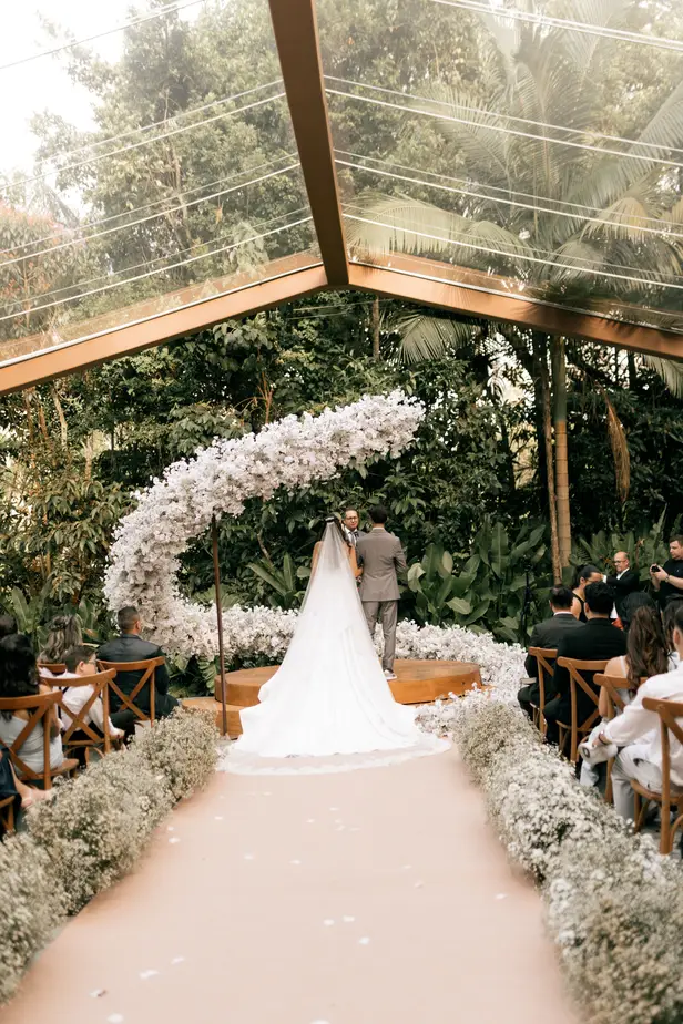 A couple beneath a white floral arch during a glasshouse ceremony